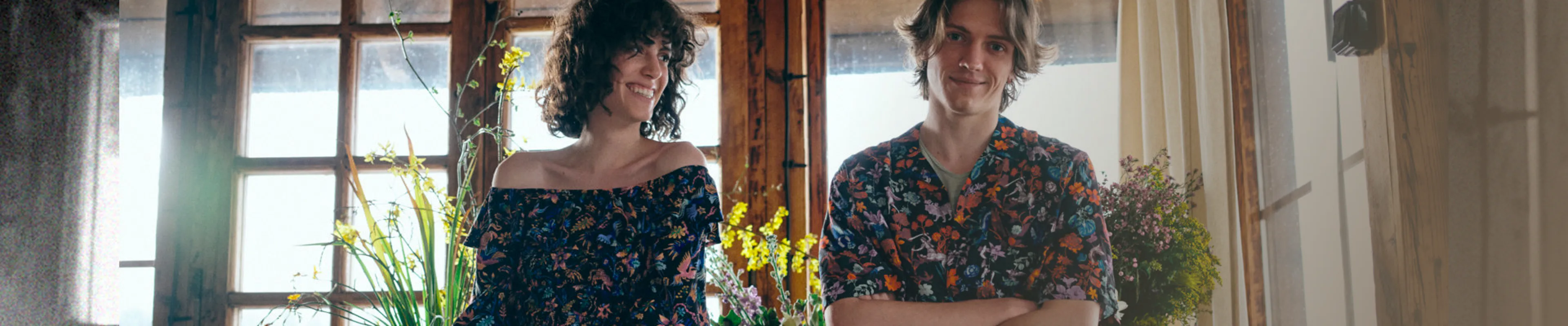 A smiling couple in colorful floral clothing sitting by a bright window.